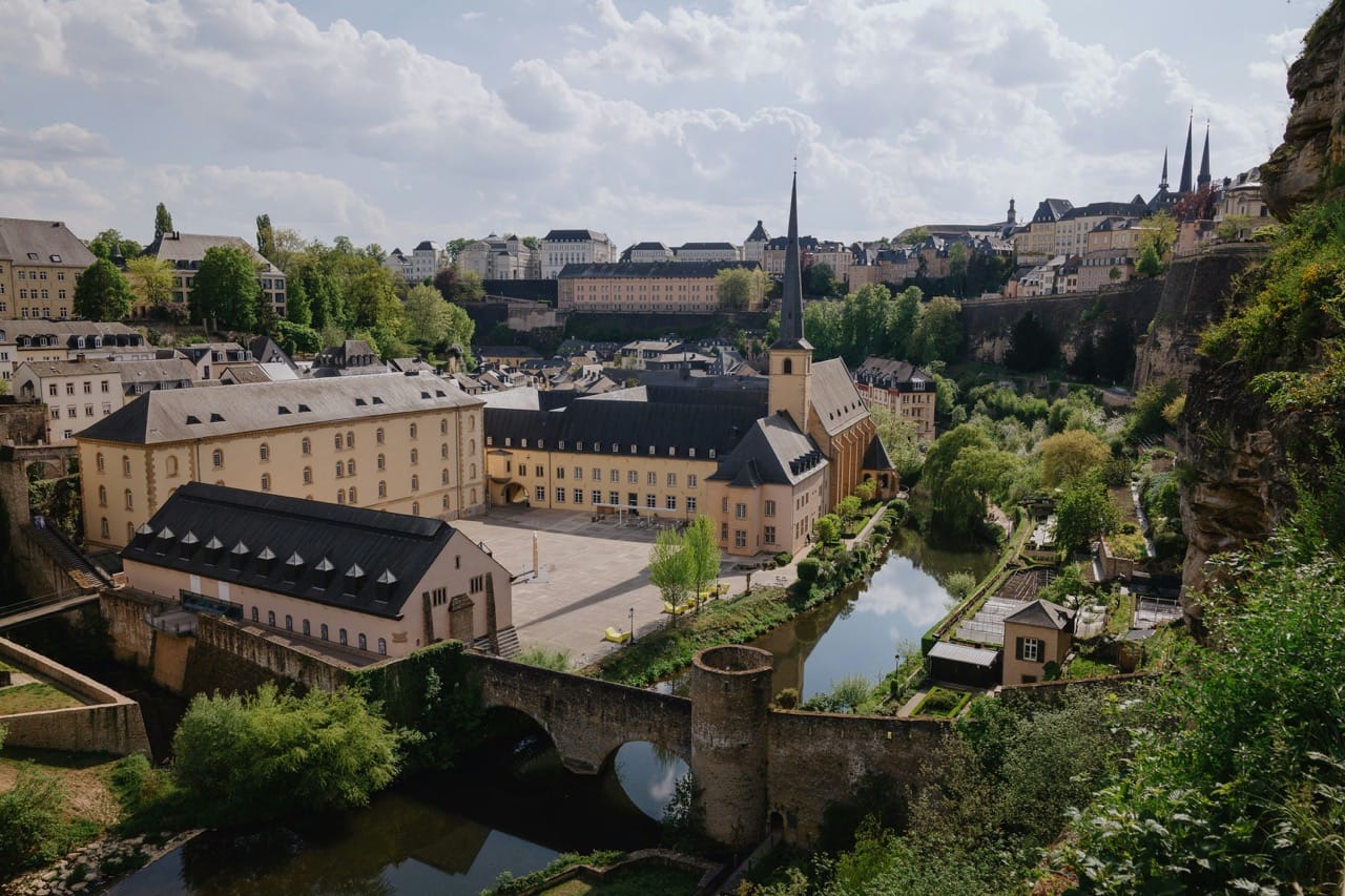 Vue panoramique du quartier historique du Grund à Luxembourg, avec l'abbaye de Neumünster, des bâtiments anciens, un pont en pierre et des espaces verts luxuriants, sous un ciel partiellement nuageux.