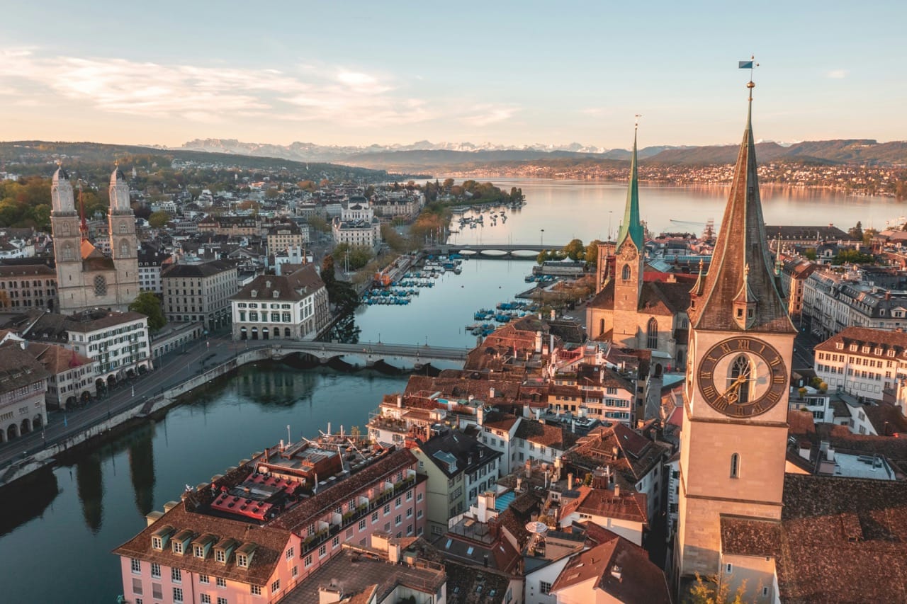 Vue aérienne de Zurich, Suisse, avec les clochers emblématiques de l'église Saint-Pierre et de la Fraumünster, le lac de Zurich en arrière-plan, et les Alpes visibles à l'horizon sous une lumière douce du matin.