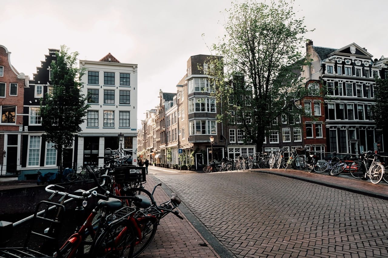 Pont typique d'Amsterdam, Pays-Bas, bordé de vélos et entouré de maisons traditionnelles aux façades étroites et aux grandes fenêtres, sous une lumière douce en fin de journée.