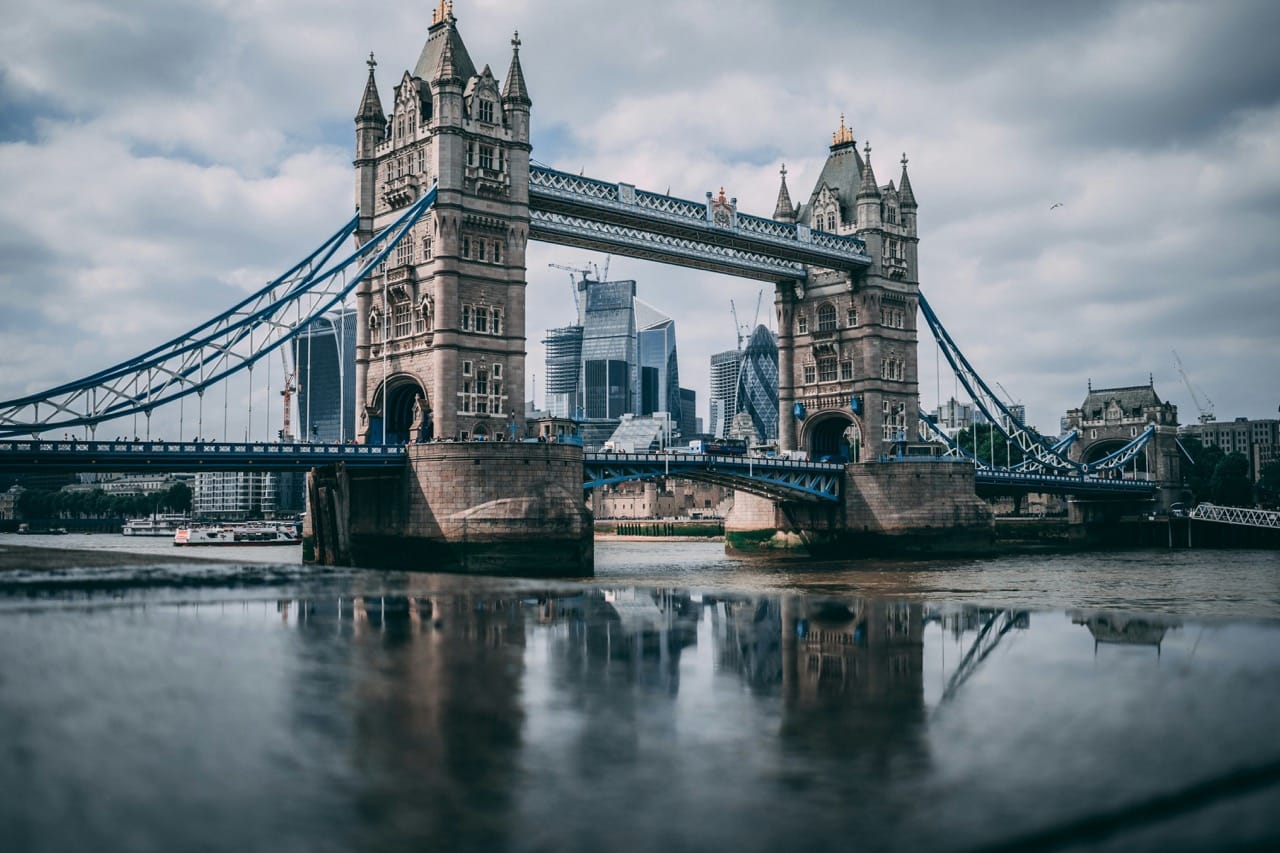 Vue emblématique du Tower Bridge à Londres, Royaume-Uni, avec des gratte-ciel modernes de la City en arrière-plan, reflétés dans les eaux de la Tamise sous un ciel nuageux.