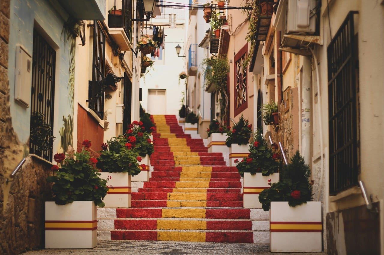 Ruelle colorée en Espagne avec des escaliers peints aux couleurs rouge et jaune du drapeau espagnol, bordés de fleurs rouges et de maisons traditionnelles ornées de balcons.