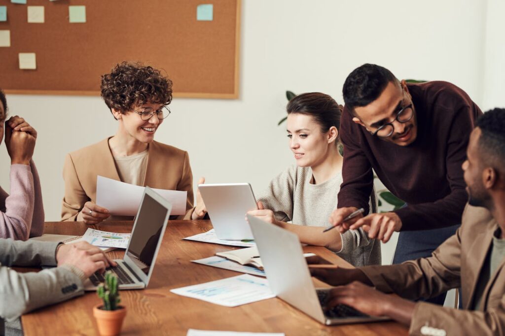 Réunion de travail collaborative entre collègues autour d’une table, avec ordinateurs, tablettes et documents. L’ambiance est détendue et productive.