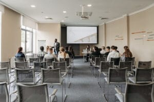 Conférence ou session de sensibilisation en entreprise, avec un intervenant debout devant un petit groupe de participants, dans une salle équipée d’un vidéoprojecteur et d’un écran de présentation.