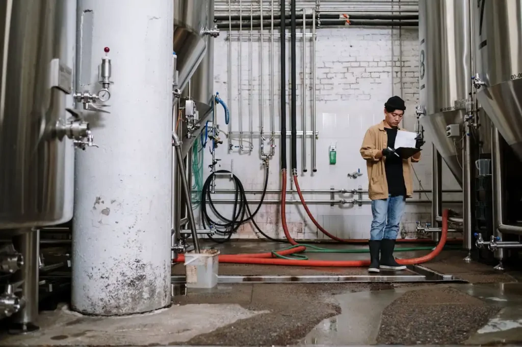 Technicien debout entre deux cuves industrielles, consultant un clipboard pour vérifier les relevés, dans un environnement de production.