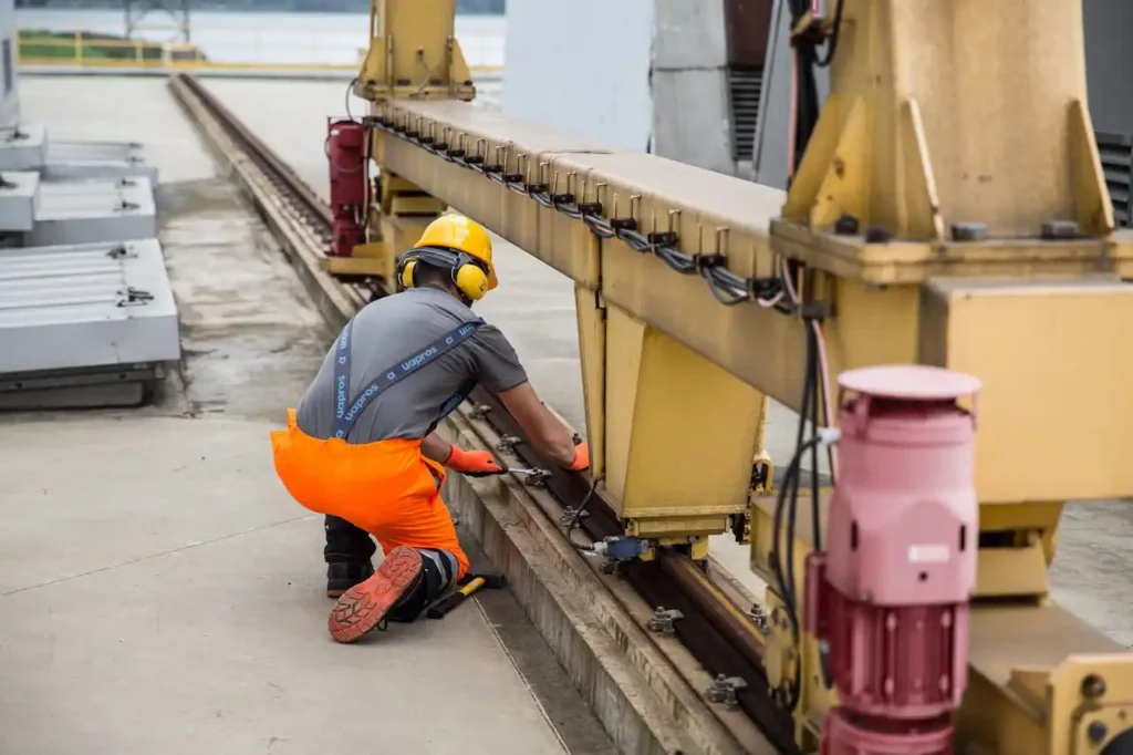 Ouvrier en tenue haute visibilité et casque de protection effectuant la maintenance d’une structure métallique sur rail dans un environnement industriel.