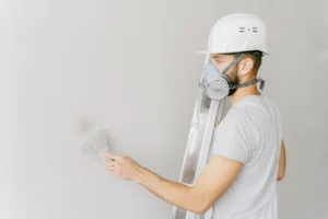 Ouvrier portant un casque et un masque de protection, utilisant une spatule pour lisser un mur.