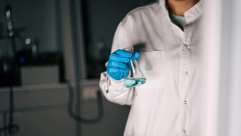 Technicien de laboratoire tenant un erlenmeyer contenant une solution liquide, équipé de gants de protection et d’une blouse.