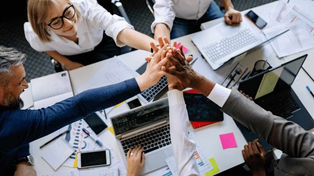 Groupe de collègues en réunion autour d’une table de travail, empilant leurs mains au centre en signe de collaboration et d’esprit d’équipe, avec des ordinateurs portables et des documents visible