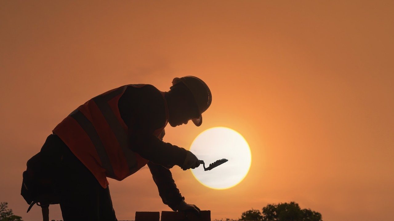Ouvrier du bâtiment travaillant au coucher du soleil avec gilet de sécurité et casque — image illustrant le travail en extérieur, l'exposition à la chaleur et la sécurité des travailleurs isolés