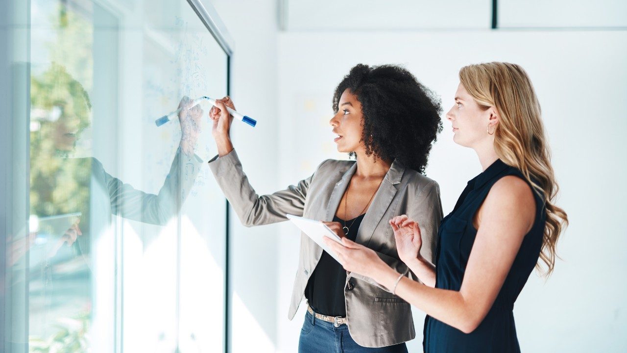 "Deux professionnelles collaborant sur un brainstorming en entreprise, écrivant sur un tableau vitré, image illustrant le travail d'équipe, la stratégie et le management en environnement de bureau moderne
