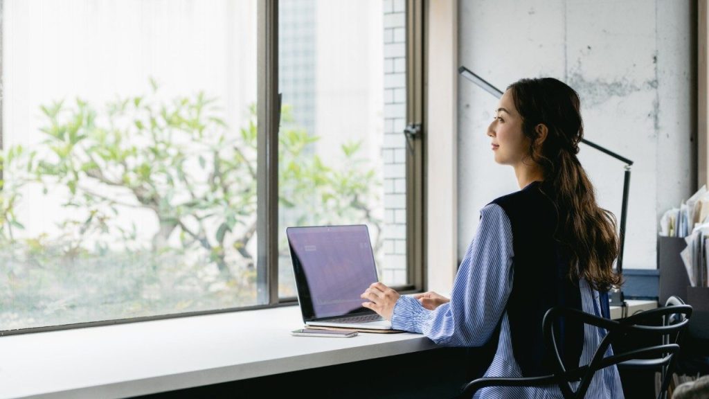 Femme travaillant sur ordinateur portable dans un bureau lumineux, réfléchissant face à la fenêtre — image représentant le télétravail, la productivité et le bien-être au travail en environnement moderne