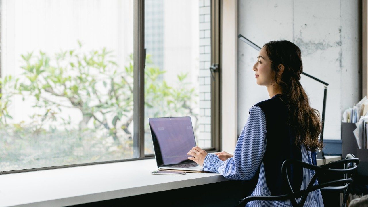 Femme travaillant sur ordinateur portable dans un bureau lumineux, réfléchissant face à la fenêtre — image représentant le télétravail, la productivité et le bien-être au travail en environnement moderne