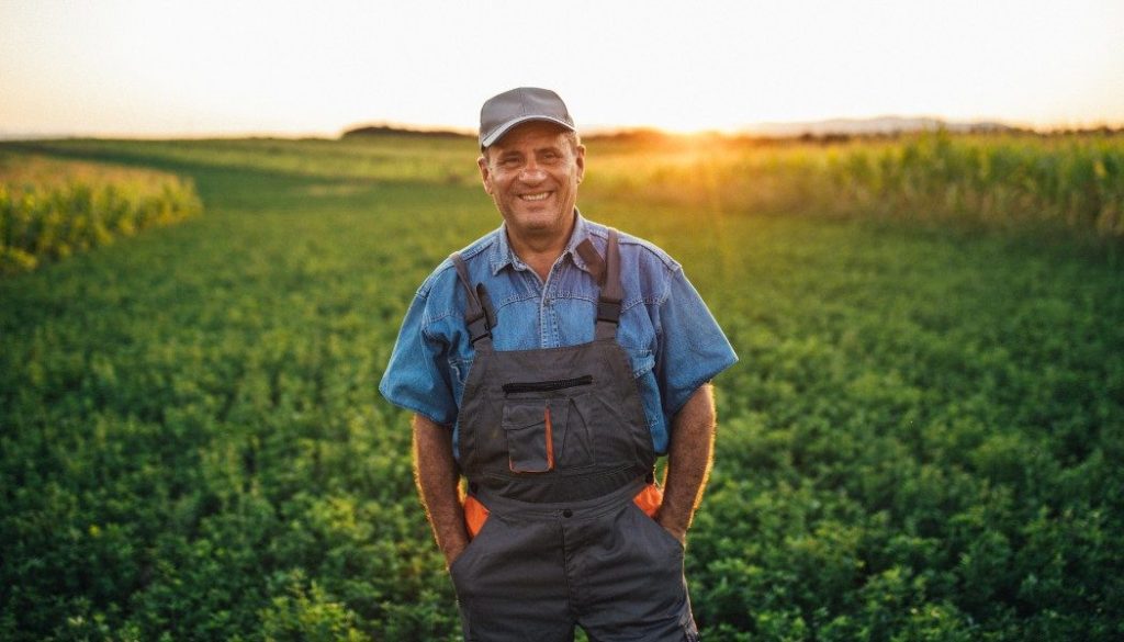 Agriculteur souriant dans son champ au coucher du soleil, illustrant les métiers agricoles exposés à l'isolement professionnel.
