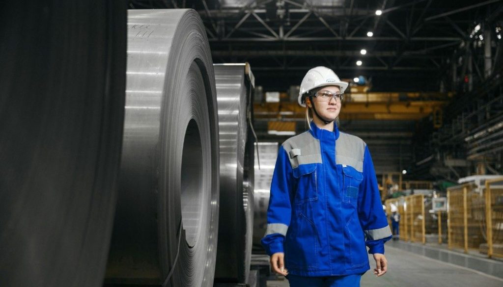 Femme ingénieure en usine sidérurgique, équipement de protection individuelle, bobines d'acier laminé, environnement industriel lourd