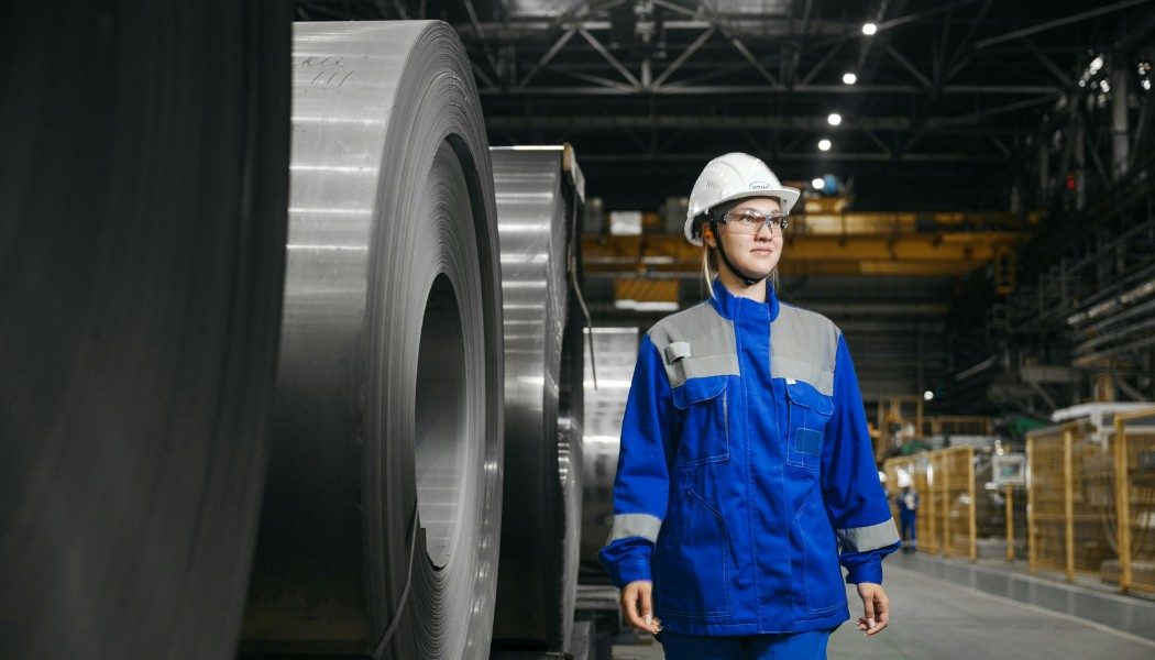 Femme ingénieure en usine sidérurgique, équipement de protection individuelle, bobines d'acier laminé, environnement industriel lourd